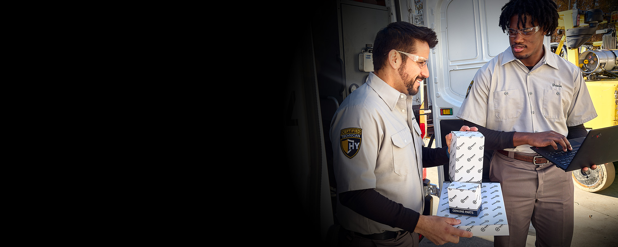 A technician carries genuine parts from his truck while another takes notes on a laptop